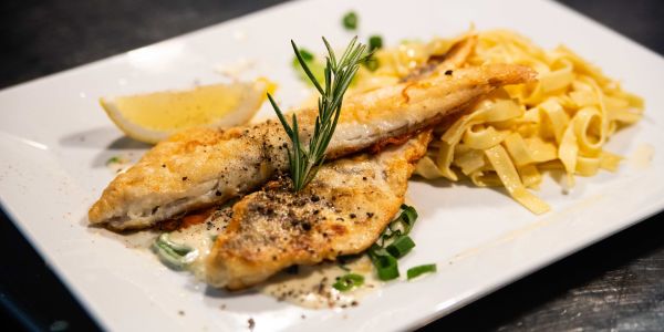 Knusprig angebratenes Fisch Filet mit Rosmarin, Pfefferkörnern und Zitrone auf Tagliatelle Pasta angerichtet
