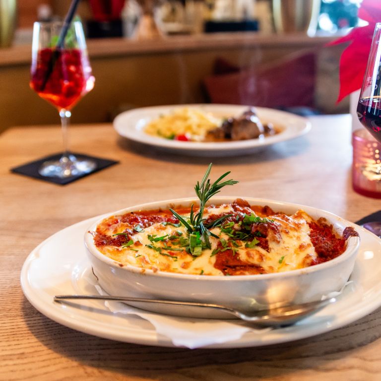 Überbackene Pasta mit Rotwein. Im Hintergrund ein weiteres Gericht und ein Cocktail.