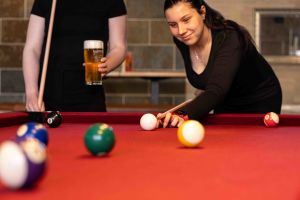 Eine junge Frau zielt konzentriert mit dem Queue beim Billardspielen, während im Hintergrund jemand ein Bierglas hält.