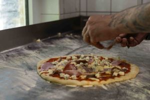 Ein Koch verteilt in der Küche mit den Händen frische Zutaten wie Schinken und Blauschimmelkäse auf einem Pizzateig.