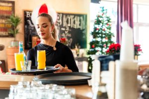 Kellnerin serviert Getränke auf einem Tablett vor weihnachtlichem Hintergrund.
