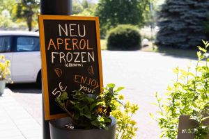 Eine schwarze Kreidetafel wirbt für „Neu Aperol Frozen (Granita)“ für 5€. Sie steht im Freien an einer Topfpflanze.