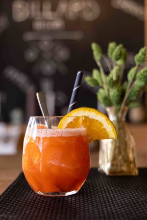 Ein leuchtend orangefarbener Cocktail in einem bauchigen Glas, dekoriert mit einer Orangenscheibe und einem schwarz-weißen Strohhalm.