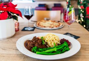 Teller mit Rinderbraten, Spätzle und grünem Spargelbrokkoli, dahinter Pizza.
