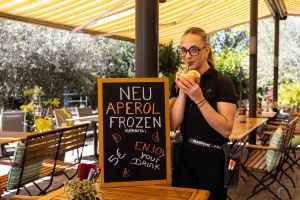 Eine lächelnde Kellnerin trinkt mit einem Strohhalm aus einem Cocktail, während sie hinter der Kreidetafel für Aperol Frozen steht.