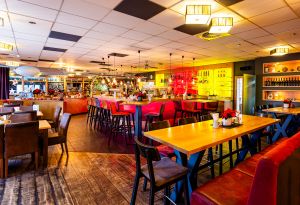 Großer Gastraum mit Holztischen, roten Bänken und beleuchteter Bar.
