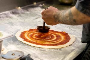 Ein Pizzabäcker verstreicht mit eine r Kelle in einer schwungvollen Spirale rote Tomatensoße auf einem ausgerollten Pizzateig.