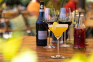 Ein leuchtend gelber Cocktail mit Zuckerrand und Zitronenscheibe, arrangiert auf einem Holztisch mit Weinflaschen im unscharfen Hintergrund.