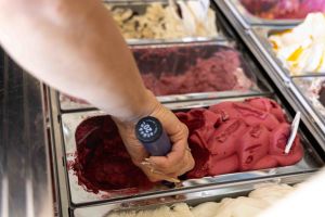 Eine Mitarbeiterin portioniert mit einem Eisportionierer tiefrotes Beeren-Sorbet aus einer metallenen Eiswanne in der Theke.