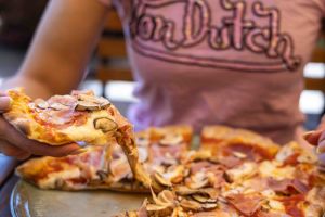 Eine Hand hebt ein Stück ofenfrische Pizza an, wobei der geschmolzene Käse Fäden zieht. Die Pizza ist mit Schinken und Pilzen belegt.