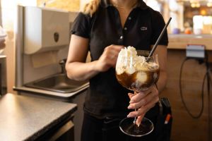 Eine Mitarbeiterin in schwarzem Poloshirt hält ein fertiges Affogato-Dessert mit Eis, Kaffee und einer großzügigen Sahnehaube.