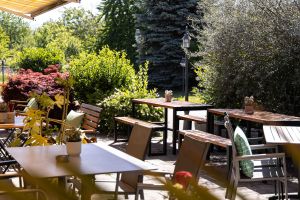 Sonnige Restaurantterrasse mit Holztischen und passenden Stühlen, umgeben von Grünpflanzen.
