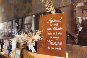 Ein gerostetes Metallschild mit ausgestanzten Formen und handgeschriebenem weißen Text über Weintherapie. Ein Trockenblumenstrauß steht daneben.