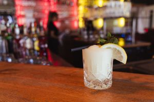 CLimoncello-Cocktail mit schwarzem Strohhalm, Zitronenscheibe und Minze auf einer Holztheke. Im unscharfen Hintergrund ist eine Bar zu sehen.