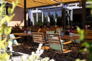 Der Außenbereich des Restaurants mit leeren Holztischen und Stühlen, die mit hellgrünen Kissen ausgestattet sind.