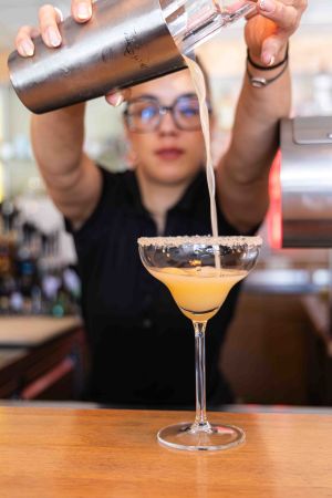 Eine Barkeeperin gießt einen frisch gemixten Cocktail aus einem metallenen Shaker in ein Glas mit Zuckerrand.