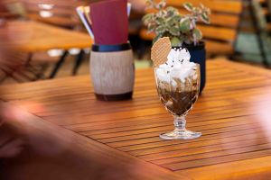 Ein dekoratives Dessertglas mit Affogato, gekrönt von einem Sahneberg, Schokostreuseln und einer Waffel, auf einem Holztisch der Terrasse.