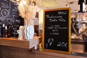 Kreidetafel auf einer Theke neben einer weißen Vase und Weinflaschen. Darauf steht ein Angebot für 2 Apero Spritz und 1 Pinsa für 20€.