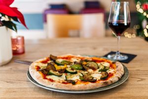 Vegetarische Pizza mit Grillgemüse neben einem Glas Rotwein auf einem Holztisch.
