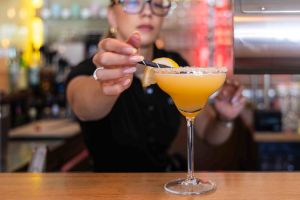 Eine Barkeeperin, die einen gelb en Cocktail im langstieligen Glas sorgfältig mit einer halben Zitronenscheibe garniert.