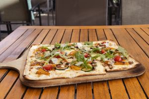 Rechteckige Pinsa belegt mit Zucchini, Tomaten, Rucola, Pilzen, Paprika und Parmesanspähnen auf einem Holzbrett, das auf einem Holztisch mit Rillen steht.