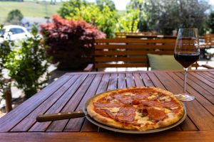 Salami-Pizza und ein Glas Rotwein auf einem Holztisch im Außenbereich in sonniger Kulisse mit Pflanzen.