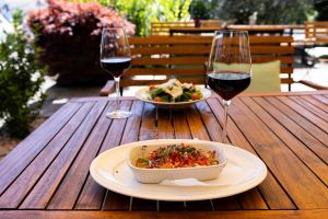 Wißer Käse in einer weißen Keramikform, belegt mit frischen Tomatenwürfeln, Zwiebeln und Kräutern. Die Form steht auf einem Terrassentisch aus Holz. Im Hintergrund ist ein Glas Rotwein und ein Saalt zu sehen.