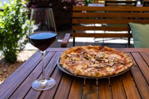 Pizza mit Schinken und frischen Champignons auf dem Terrassentisch, begleitet von einem Glas Rotwein.