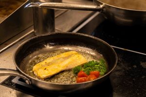 Ein helles Fischfilet brät in einer Pfanne in schäumender Butter, garniert mit Kirschtomaten und frischer Petersilie.