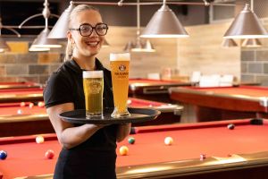 Eine lächelnde Kellnerin präsentiert ein Tablett mit zwei frisch gezapften Bieren inmitten eines Raums mit mehreren Billardtischen.