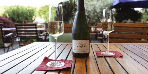 Eine Sektflasche vom Weingut Engler und zwei gefüllte Sektgläser stehen auf quadratischen roten Servietten auf einem Holztisch im Freien.