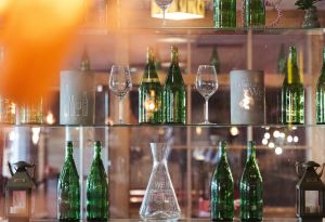 Zwei Glasregale mit grünen Weinflaschen, Weingläsern und runden Deko-Zylindern mit gravierten Motiven, darunter einer mit Text über Wein.