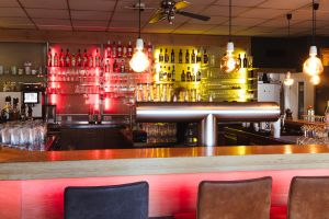 Thekenbereich einer Bar mit metallener Zapfanlage im Vordergrund. Dahinter stehen beleuchtete Regale mit Flaschen und Gläsern. Die Beleuchtung ist rot und gelb.
