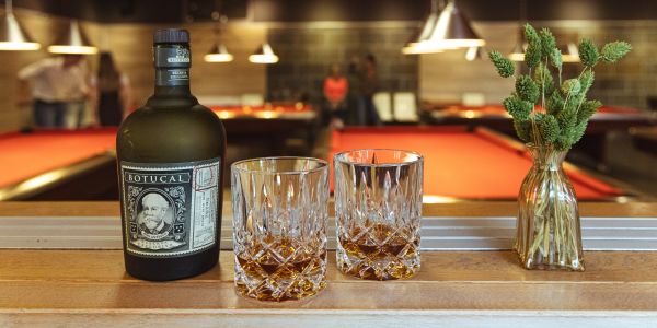 Eine Flasche Botucal Rum und zwei Kristallgläser mit Rum auf einer Holztheke. Im unscharfen Hintergrund stehen Personen an roten Billardtischen.