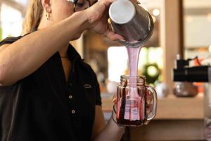 Ein cremiger, hellrosa Milchshake fließt aus einem Shaker in ein bauchiges Henkelglas, dessen Rand mit Schokoladensoße verziert ist.
