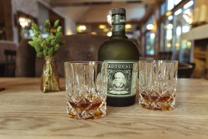 Eine Flasche Botucal Rum und zwei Kristallgläser mit Rum auf einem Holztisch. Eine kleine Vase mit getrockneten Gräsern steht links daneben.