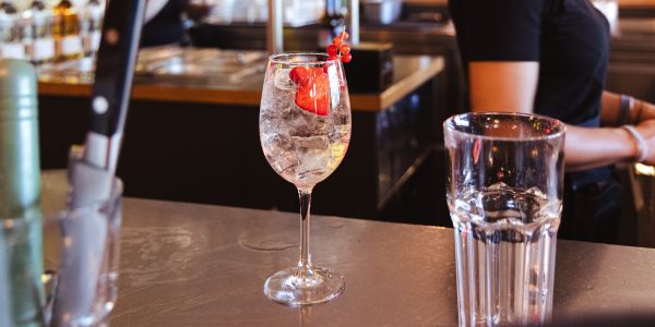 Ein Stielglas mit einem klaren Cocktail, Eis, Erdbeeren und roten Johannisbeeren als Garnitur steht auf einer Bar.