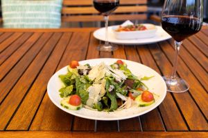 Ein frischer Rucola-Salat mit halbierten Kirschtomaten, Gurkenscheiben und üppigen Parmesanspänen auf einem weißen Teller im Außenbereich.