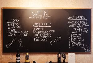 Große schwarze Kreidetafel an einer Wand mit einer handgeschriebenen Weinkarte, unterteilt in offene Rot-, Weiß- und Roséweine. Davor stehen Weinflaschen und Gläser.