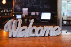 Ein massiver Dekoschriftzug mit dem Wort "Welcome" aus hellem Material steht auf einer Holztheke vor einem unscharfen Bar-Hintergrund.