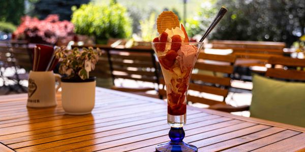 Ein großer, fruchtiger Eisbecher mit Erdbeeren, Sahne und einer Eiswaffel in einem hohen, blauen Glas auf einem Holztisch.