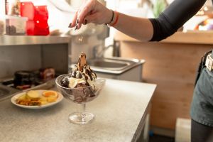 Eine Hand streut Krokant-Topping über einen Schokoladen-Eisbecher.
