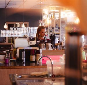 Eine Barkeeperin arbeitet hinter einer gut beleuchteten Bar mit Zapfanlage. Auf der Theke stehen unterschiedliche Bar-Utensilien.