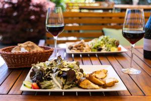 Zwei Teller mit frischem Salat und saftigen Hähnchenbruststreifen auf einem Holztisch im Freien, samt einem Brotkorb und Rotwein.