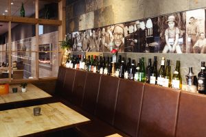 Eine lange, braune Lederbank und Holztische in einem Restaurant. Eine Reihe von Weinflaschen mit Lichterkette und historische Fotos zieren die Wand darüber.