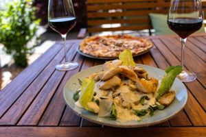 Salat mit gegrillten Hähnchenstreifen, Parmesanspänen und Dressing auf einem Holztisch, im Hintergrund eine Pizza und Wein.