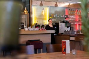 Eine freundliche Mitarbeiterin steht hinter der hölzernen Bar, auf der ein dekorativer "Welcome"-Schriftzug platziert ist.