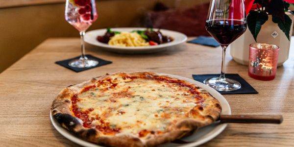 Pizza auf einem weißen Teller auf einem Holztisch im Restaurant; im Hintergrund eine weitere Speise und ein Getränk.