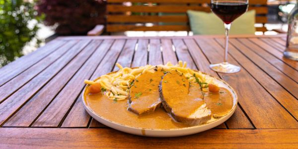 Zwei Scheiben Schweinebraten mit reichlich Soße und schwäbischen Spätzle auf einem Holztisch, im Hintergrund ein Glas Rotwein.