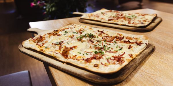 Zwei Flammkuchen belegt mit Speck, Zwiebeln und Kräutern auf Holzbrettern auf einem Holztisch, einer näher im Vordergrund, einer weiter hinten.